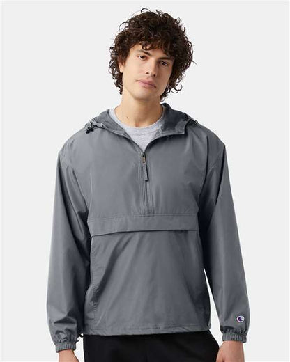 A young man with curly hair wears the Champion CO200, a wind- and water-resistant micro poplin jacket with a front zipper and large front pocket, standing facing forward against a plain light gray background.