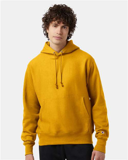 A young person with curly brown hair wears a mustard yellow Champion S101 hoodie featuring a front pocket and drawstrings, standing against a plain light gray background.