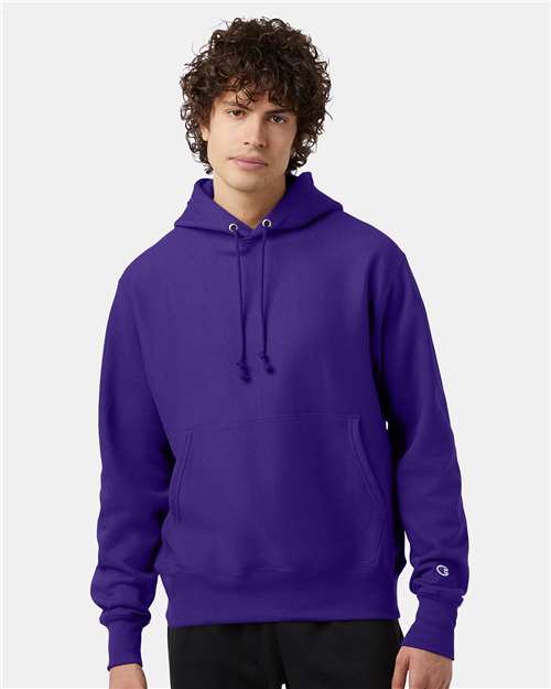 A young person with curly brown hair wears a plain purple Champion S101 sweatshirt featuring a front pocket and drawstrings, standing against a light gray background.