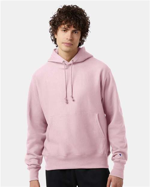 A young person with curly dark hair wears the Champion S101 Reverse Weave hoodie in light pink, featuring a front pocket and hood strings, standing against a plain light gray background.