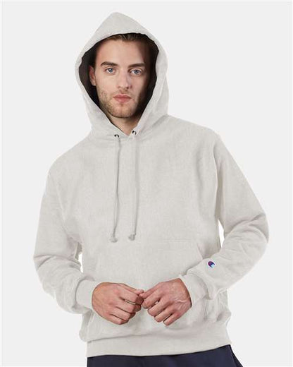 A person wears a light gray Champion S101 hoodie with the hood up, looking at the camera with a neutral expression and hands lightly touching. The Champion S101 offers comfort, style, and is recognized for its sustainable manufacturing.