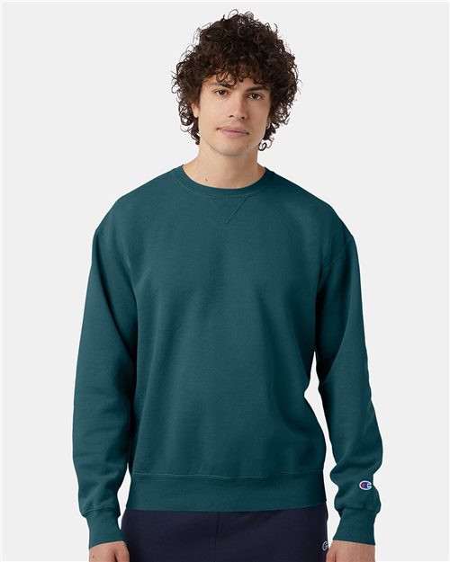 A young man with curly hair wears the Champion CD400 no-shrink cotton teal sweatshirt and dark pants, standing against a plain light background.