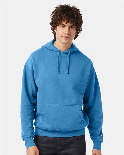 A young man with curly hair wears a bright blue Champion CD450 hoodie, made from no-shrink cotton with a front pocket and drawstrings, standing against a plain light backdrop.