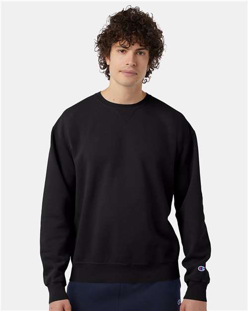 A young man with curly hair wears the Champion CD400 sweatshirt in black, made with no-shrink cotton and paired with dark pants. The Champion logo appears on the sleeve as he stands against a light gray background.