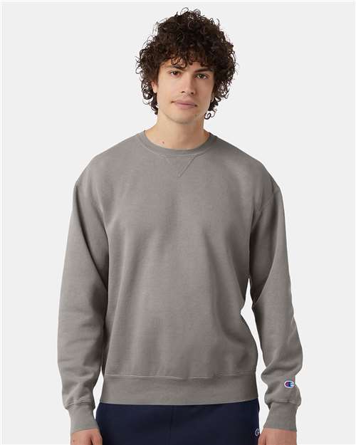 A young man with curly hair wears a Champion CD400 plain gray sweatshirt made from no-shrink cotton and dark pants, standing against a plain white background.