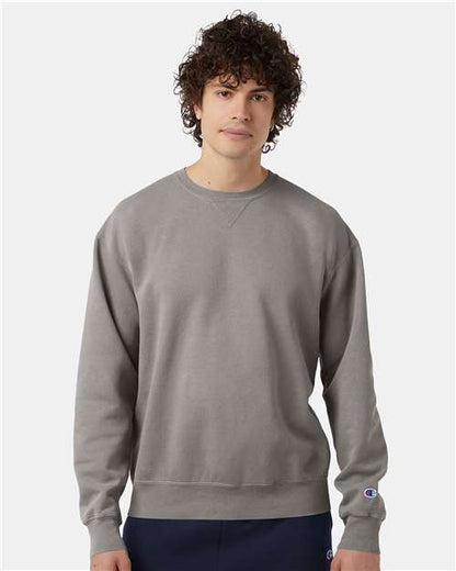 A young man with curly hair wears a Champion CD400 plain gray sweatshirt made from no-shrink cotton and dark pants, standing against a plain white background.