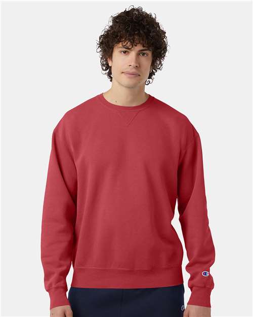 A person with curly hair wears a plain red Champion CD400 sweatshirt made of no-shrink cotton and dark pants, stands against a light background, and looks at the camera with a neutral expression.