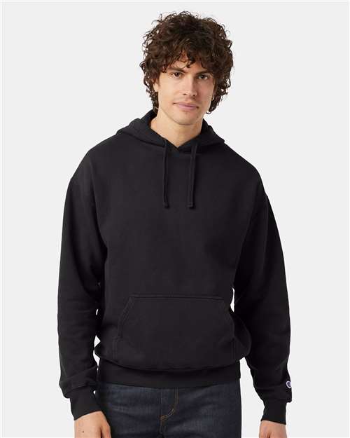 A young man with curly brown hair wears a plain black Champion CD450 hoodie made from no-shrink cotton and dark jeans, standing against a light gray background.