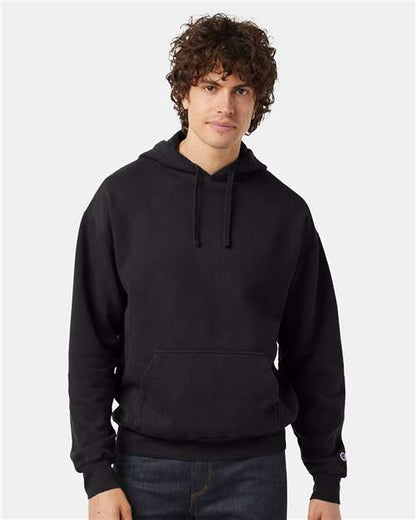 A young man with curly brown hair wears a plain black Champion CD450 hoodie made from no-shrink cotton and dark jeans, standing against a light gray background.