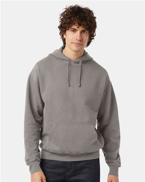 A young man with curly brown hair wears a light gray Champion CD450 hoodie made from no shrink cotton, standing against a plain white background.