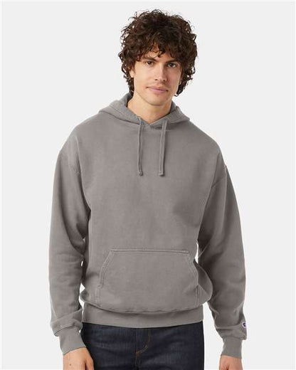 A young man with curly brown hair wears a light gray Champion CD450 hoodie made from no shrink cotton, standing against a plain white background.