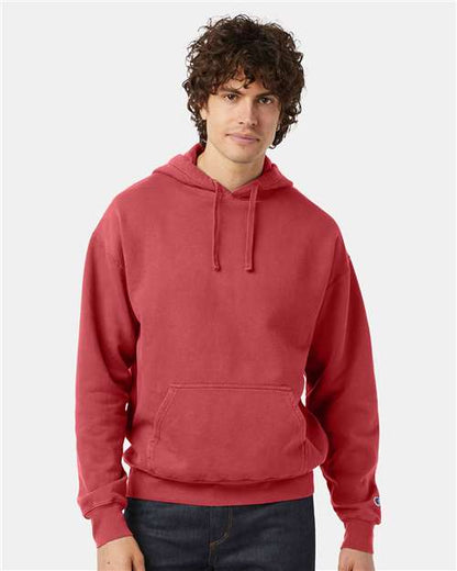 A person with curly brown hair wears a red Champion CD450 no-shrink cotton hoodie and dark jeans, standing against a plain light gray background.