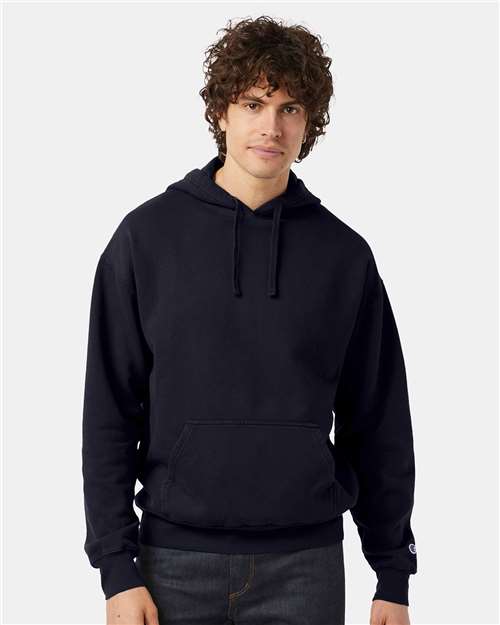 A person with curly brown hair is wearing a black Champion CD450 hoodie made from no-shrink cotton and dark jeans, standing against a plain light background and looking at the camera with a neutral expression.