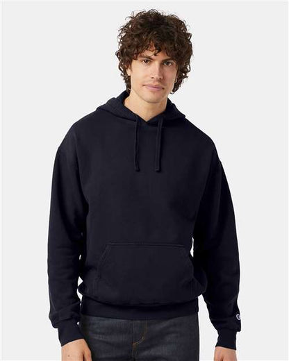 A person with curly brown hair is wearing a black Champion CD450 hoodie made from no-shrink cotton and dark jeans, standing against a plain light background and looking at the camera with a neutral expression.