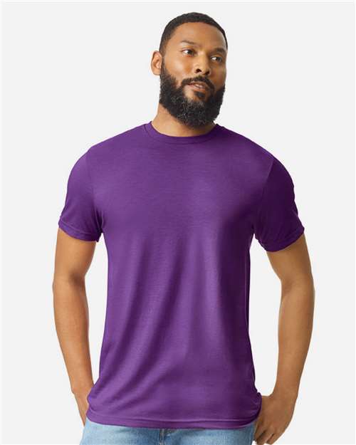 A bearded man in light blue jeans wears a Gildan Softstyle 64000CVC plain purple t-shirt, made from ring-spun cotton, standing with hands in his pockets against a light gray background.