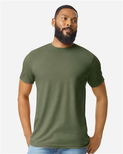 A man with a beard stands against a plain light background, wearing an olive green Gildan Softstyle 64000CVC t-shirt by Gildan made from ring-spun cotton and light blue jeans, with his hands in his pockets.