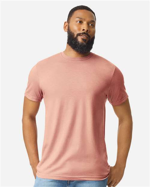 A bearded man stands against a plain light background, wearing a dusty pink Gildan Softstyle 64000CVC t-shirt made from ring-spun cotton by Gildan and light blue jeans, with his hands in his pockets.