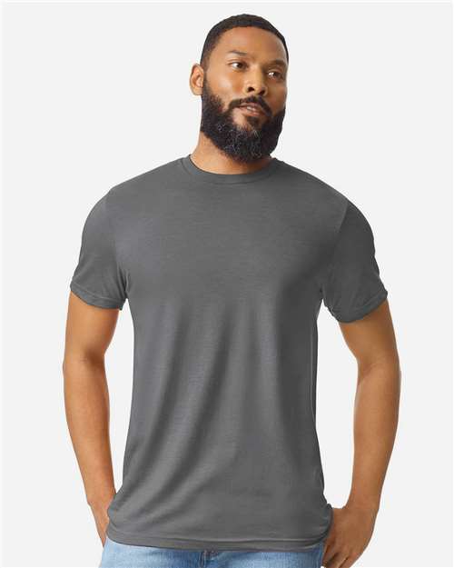 A bearded man with short hair wears a plain gray Gildan Softstyle 64000CVC t-shirt by Gildan, made from ring-spun cotton, and light blue jeans, standing with hands in his pockets against a light gray background.