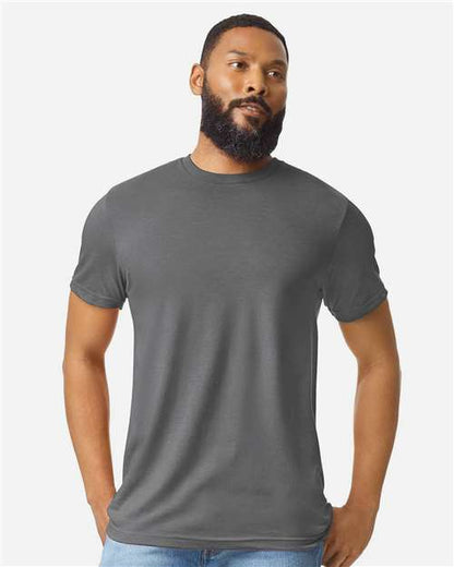 A bearded man with short hair wears a plain gray Gildan Softstyle 64000CVC t-shirt by Gildan, made from ring-spun cotton, and light blue jeans, standing with hands in his pockets against a light gray background.