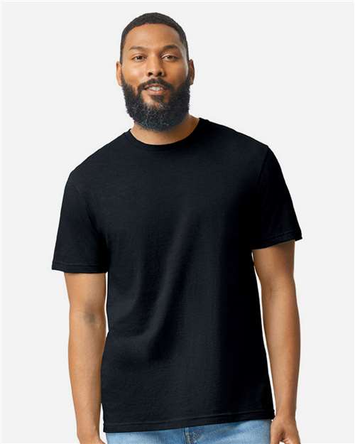 A bearded man with short hair stands facing forward, wearing a plain black Gildan Softstyle 64000CVC t-shirt by Gildan and blue jeans against a light gray background.