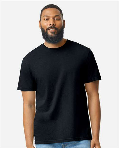 A bearded man with short hair stands facing forward, wearing a plain black Gildan Softstyle 64000CVC t-shirt by Gildan and blue jeans against a light gray background.