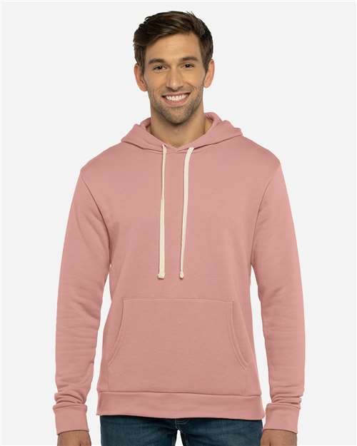A man wearing the Next Level 9303 light pink cotton-poly hoodie with a jersey-lined hood and white drawstrings stands smiling against a plain, light-gray background.