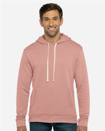 A man wearing the Next Level 9303 light pink cotton-poly hoodie with a jersey-lined hood and white drawstrings stands smiling against a plain, light-gray background.