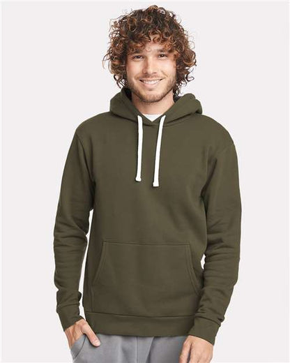 A smiling person with curly hair wears the Next Level 9303 olive green cotton-poly hoodie with a jersey-lined hood, white drawstrings, and front pocket, standing against a plain light background.