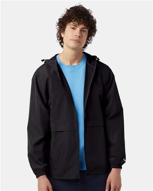 A young man with curly hair wears the Champion CO125 black wind- and water-resistant polyester micro poplin jacket over a light blue shirt, standing against a plain light gray background with a calm, neutral expression.