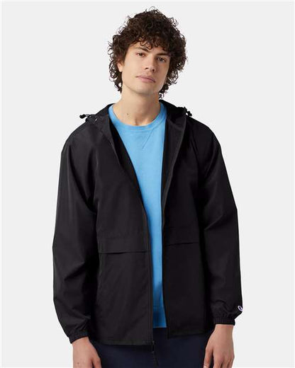 A young man with curly hair wears the Champion CO125 black wind- and water-resistant polyester micro poplin jacket over a light blue shirt, standing against a plain light gray background with a calm, neutral expression.