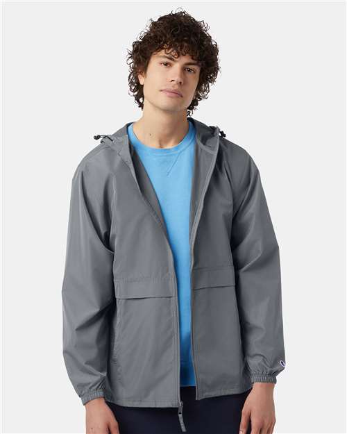 A young man with curly hair wears a light blue T-shirt and the Champion CO125 gray micro poplin wind- and water-resistant jacket, featuring a Champion patch, standing facing forward against a plain light background.