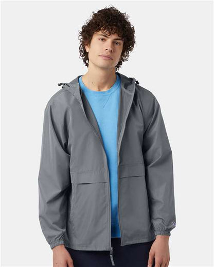 A young man with curly hair wears a light blue T-shirt and the Champion CO125 gray micro poplin wind- and water-resistant jacket, featuring a Champion patch, standing facing forward against a plain light background.