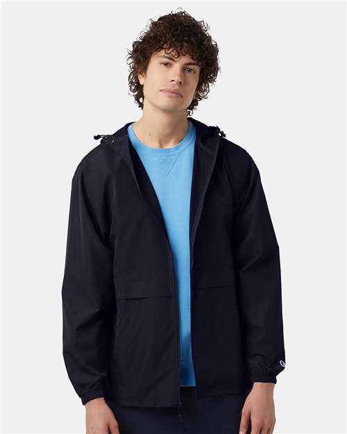 A young man with curly brown hair wears the Champion CO125 black polyester micro poplin wind and water resistant jacket over a light blue shirt, standing against a plain light gray background.