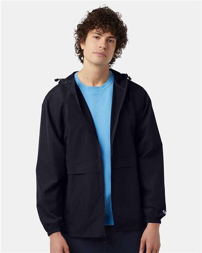 A young man with curly brown hair wears the Champion CO125 black polyester micro poplin wind and water resistant jacket over a light blue shirt, standing against a plain light gray background.