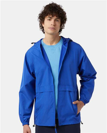 A young man with curly hair wears the Champion CO125, a bright blue polyester micro poplin wind and water resistant jacket with a Champion patch over a light blue t-shirt, standing against a plain light background.