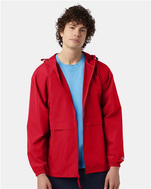 A person with curly hair wears a bright red Champion CO125 wind and water resistant jacket over a light blue shirt, standing against a plain light background and looking at the camera with a neutral expression.