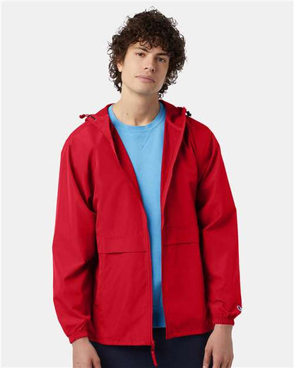 A person with curly hair wears a bright red Champion CO125 wind and water resistant jacket over a light blue shirt, standing against a plain light background and looking at the camera with a neutral expression.
