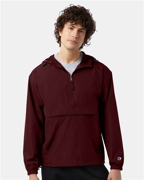 A young man with curly hair models the Champion CO200, a maroon micro poplin jacket that's wind- and water-resistant, featuring a front zipper and large pocket, shown against a plain light background.
