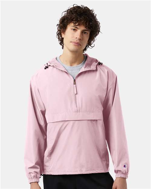A young man with curly brown hair wears the Champion CO200 light pink hooded micro poplin wind- and water-resistant jacket over a gray shirt, standing against a plain light background.