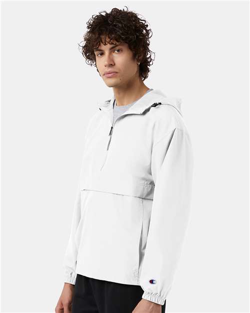 A young man with curly hair wears the Champion CO200 jacket, a wind- and water-resistant micro poplin style with a half-zip and elastic cuffs, standing against a plain light background.