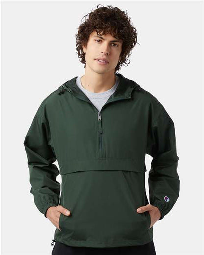 A young man with curly hair wears the Champion CO200 dark green micro poplin wind- and water-resistant half-zip jacket with a front pocket, standing with his hands in the pockets against a plain light background.