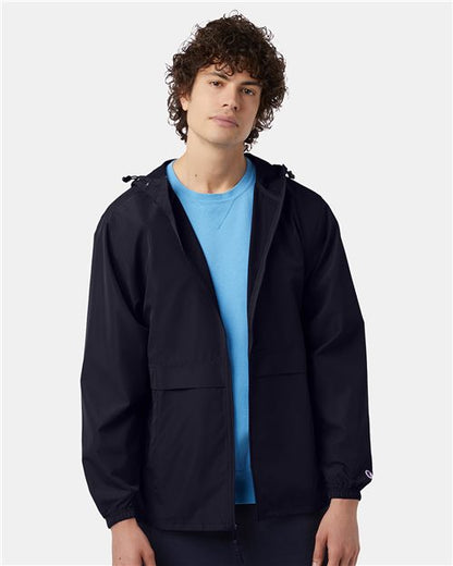 A young person with curly hair wears the Champion CO125, a black wind and water resistant jacket, over a light blue shirt, standing against a plain light gray background and looking at the camera with a neutral expression.