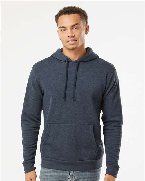A young man stands against a plain white background, wearing the Next Level 9302 dark blue cotton-polyester fleece hoodie with a front pocket and tear away label, looking at the camera with a slight smile.