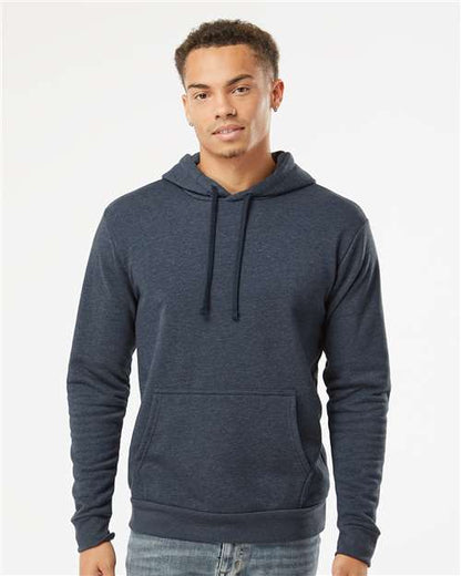 A young man stands against a plain white background, wearing the Next Level 9302 dark blue cotton-polyester fleece hoodie with a front pocket and tear away label, looking at the camera with a slight smile.