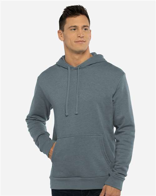 A man in the Next Level 9302 cotton-polyester fleece hoodie, blue-gray with front pocket and drawstrings, stands against a plain light background, one hand in his pocket and wearing a neutral expression.