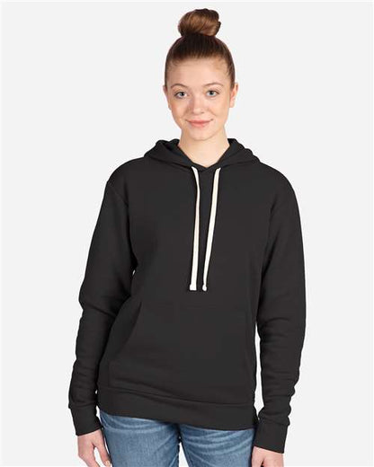 A woman with light brown hair in a bun wears the Next Level 9303 black hoodie, featuring a cotton-polyester blend, jersey-lined hood, and white drawstrings, paired with blue jeans against a plain light gray background.