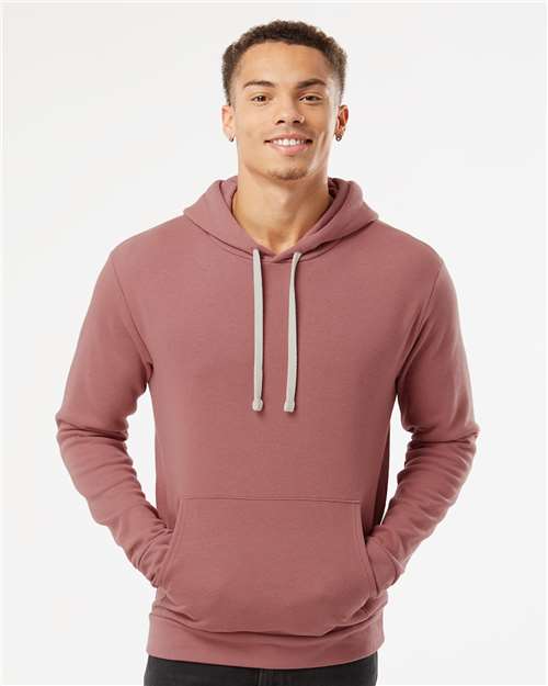 A young man smiles in a Next Level 9303 mauve cotton-polyester hoodie with a jersey-lined hood and front pocket, standing against a plain white background.