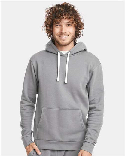 A smiling person with curly hair wears the Next Level 9303 hoodie in light gray—made of cotton polyester, featuring white drawstrings, front pocket, and jersey lined hood—standing against a plain light background.