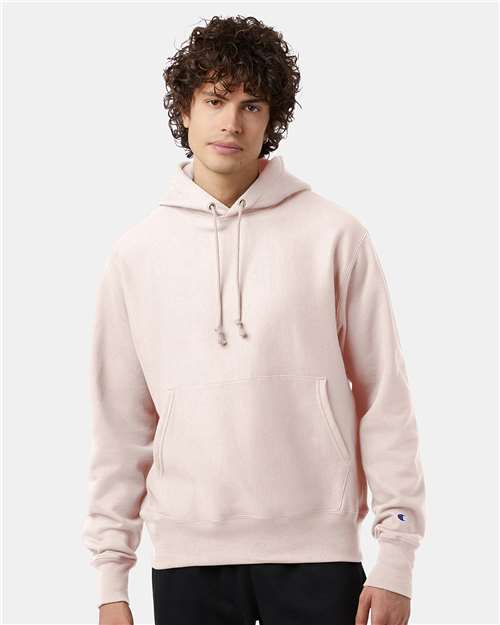 A young person with curly hair wears the Champion S101 light pink Reverse Weave hoodie, featuring a front pocket and drawstrings, against a plain gray background. This hoodie combines comfort with sustainable manufacturing.