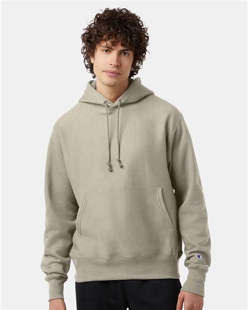 A young person with curly hair wears a light gray Champion S101 Reverse Weave hoodie with a front pocket, standing against a plain white background.